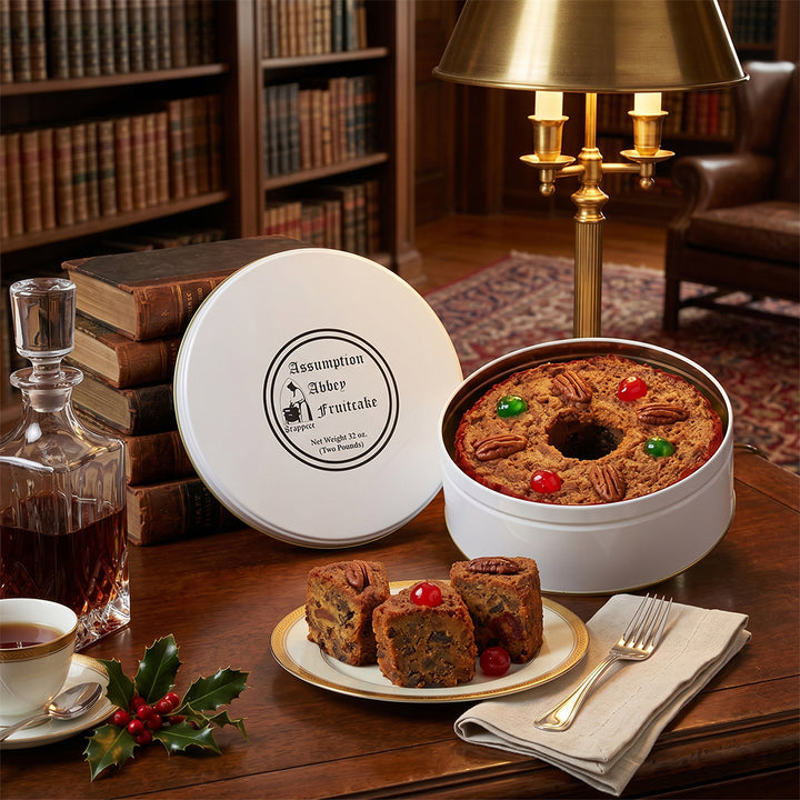 Assumption Abbey Fruitcake in a gift tin with slices on a plate, surrounded by books and a lamp in a cozy room.